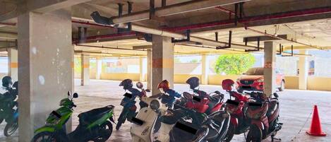 A covered parking area with multiple scooters parked, concrete pillars, and a red traffic cone.