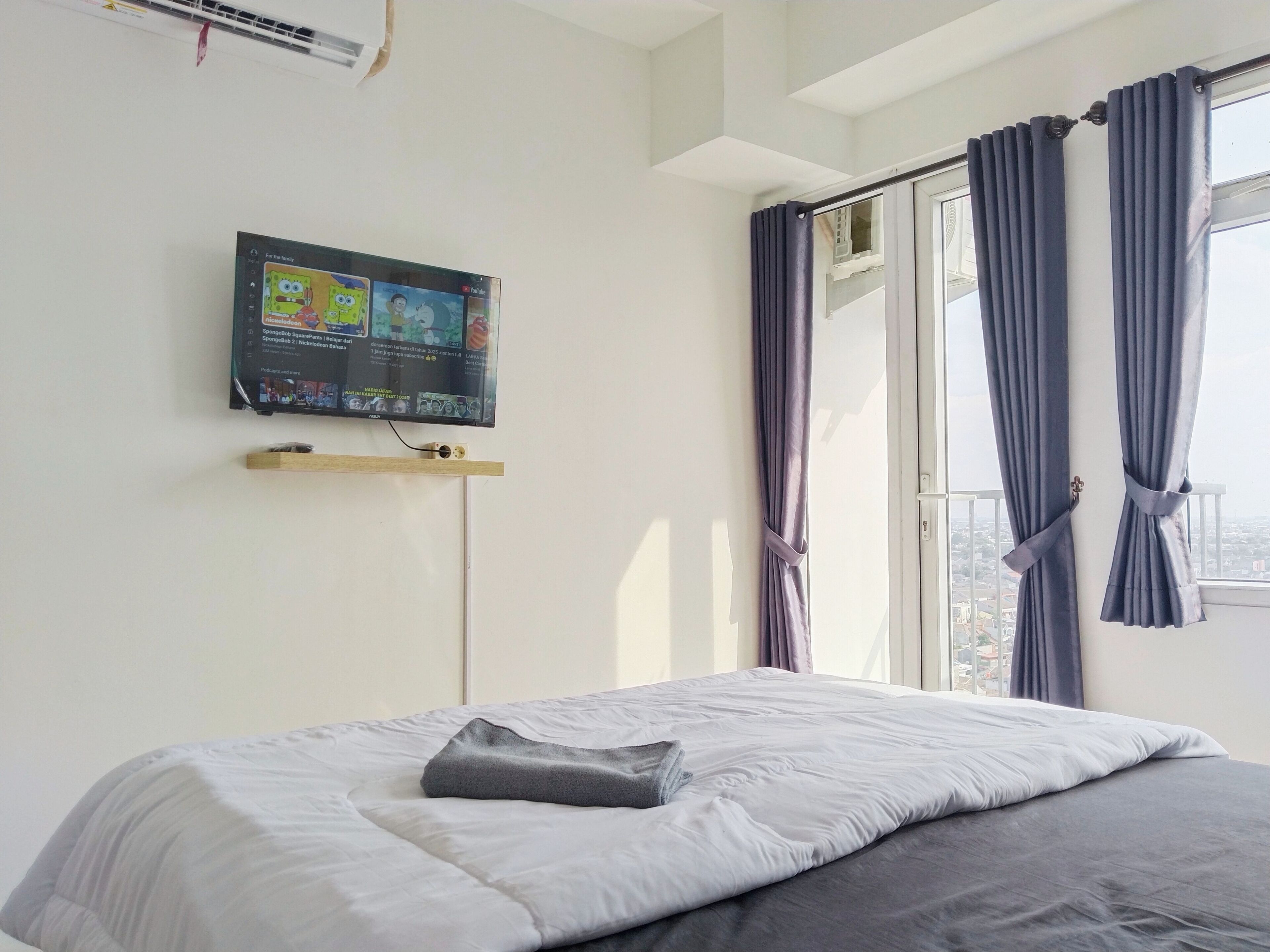 A hotel room with a bed, a flat-screen TV mounted on the wall, and a window with curtains.
