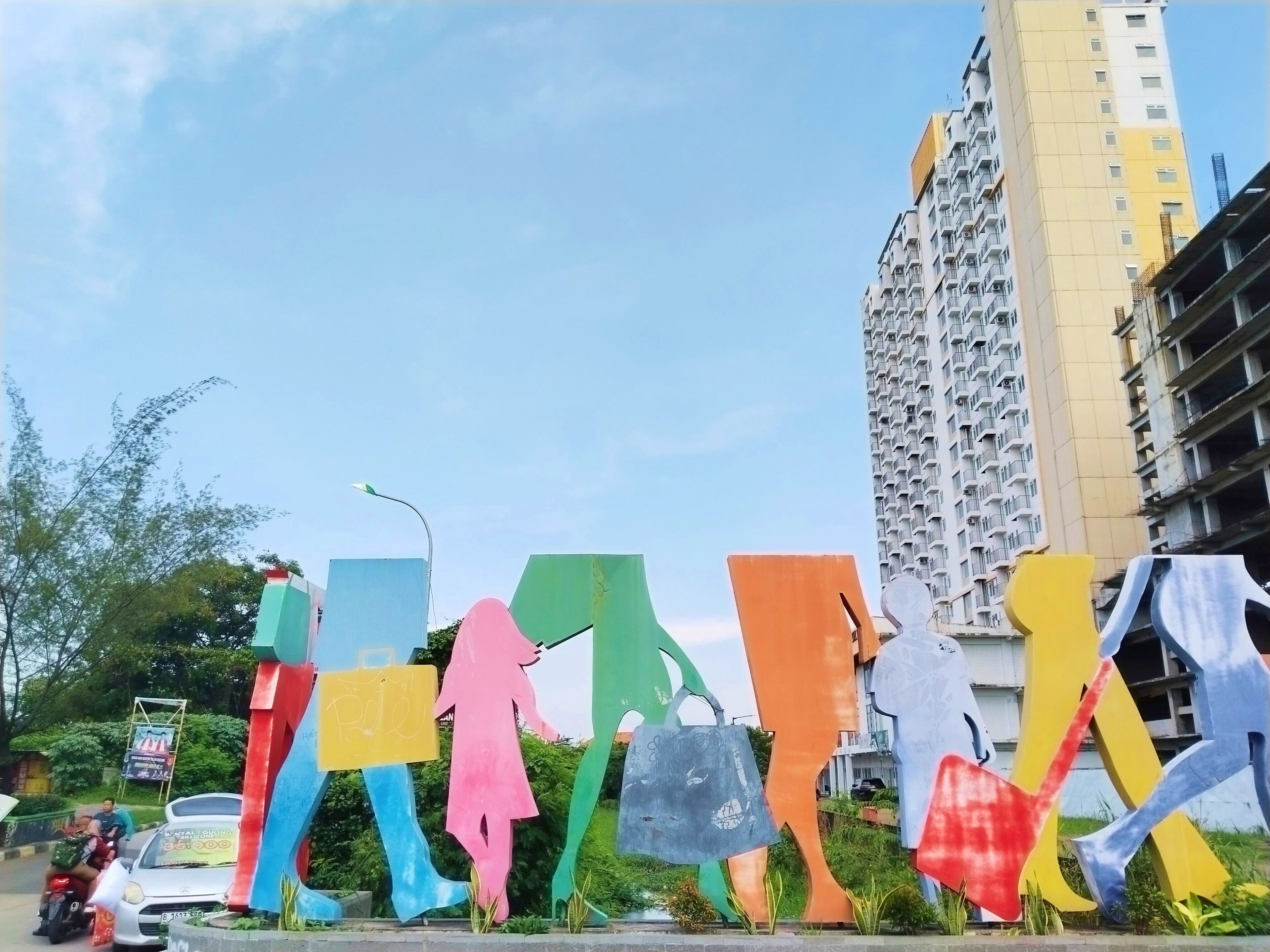 Una scultura colorata raffigurante figure umane in diverse tonalità, un palazzo residenziale imponente e una strada con veicoli e vegetazione.