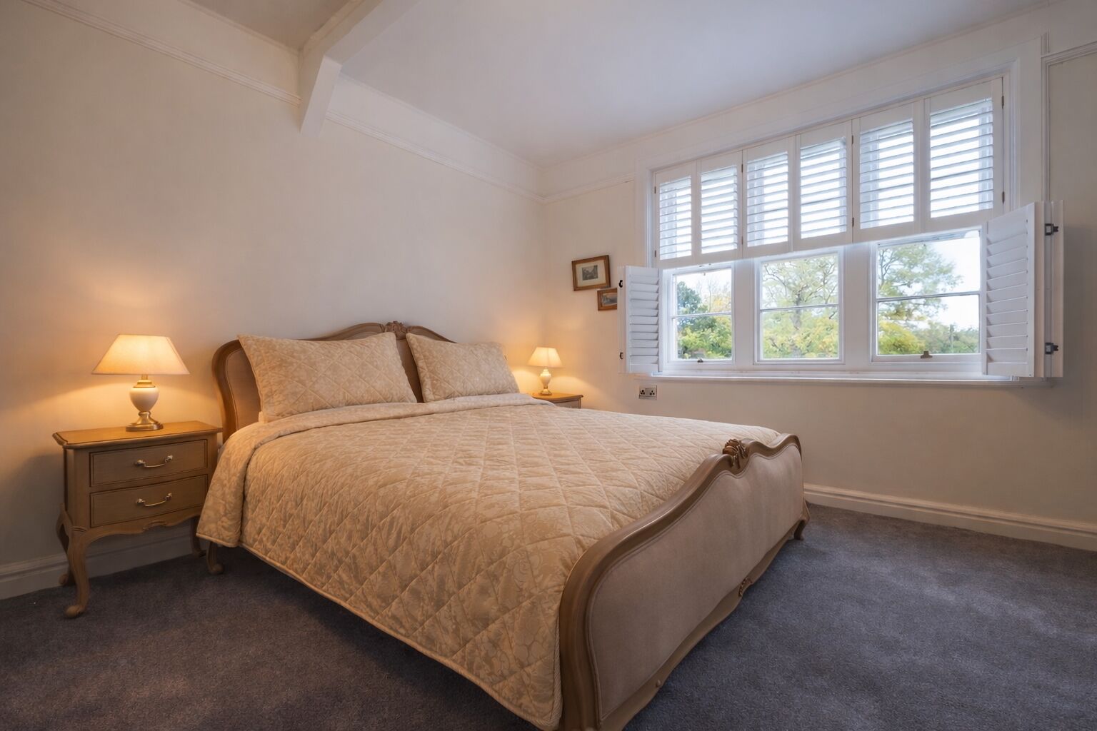 A bedroom with a bed, two bedside tables with lamps, a window with shutters, and a framed picture on the wall.