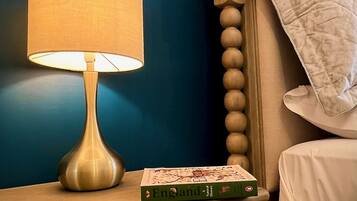 A bedside table with a lamp, a book titled 'England Natural History', and a bed with a white pillow against a blue wall.