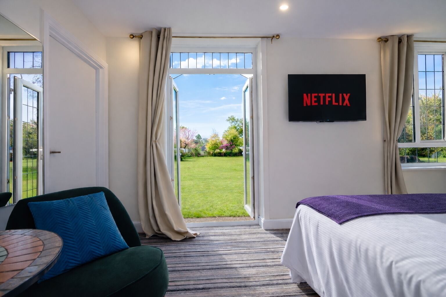 Habitación de hotel con cama, sofá verde, pantalla de televisión mostrando "NETFLIX" y vista a un jardín por una puerta abierta.