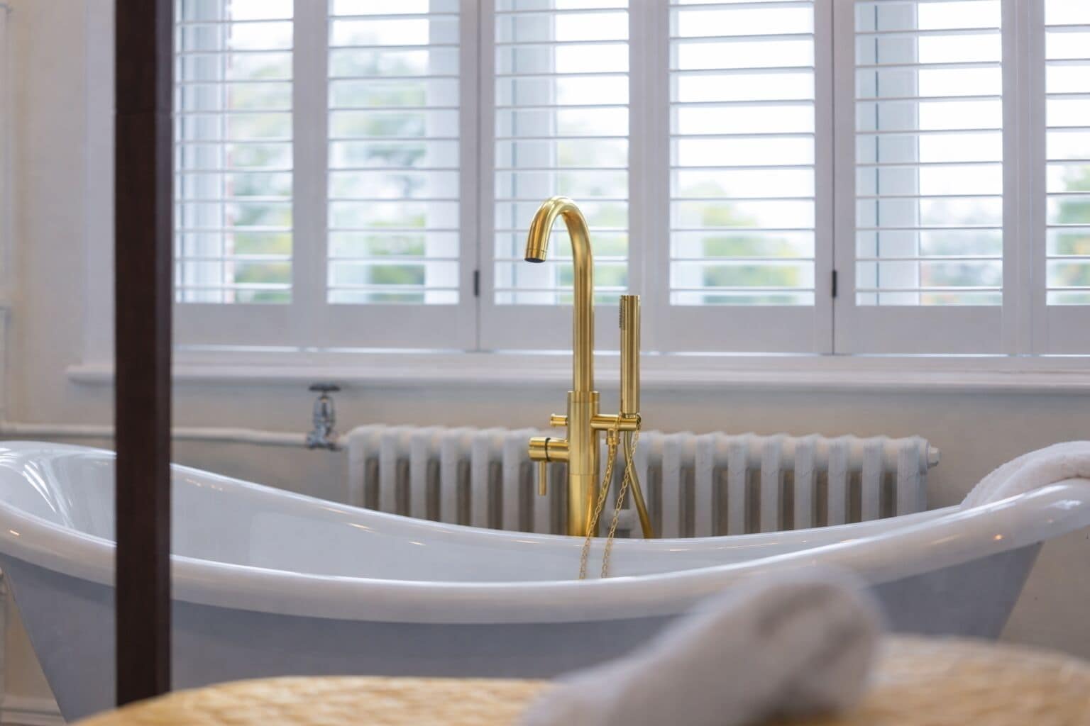A modern bathroom with a freestanding bathtub, a gold faucet, and a window with blinds.