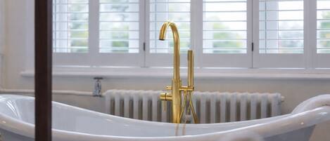 A modern bathroom with a freestanding bathtub, a gold faucet, and a window with blinds.