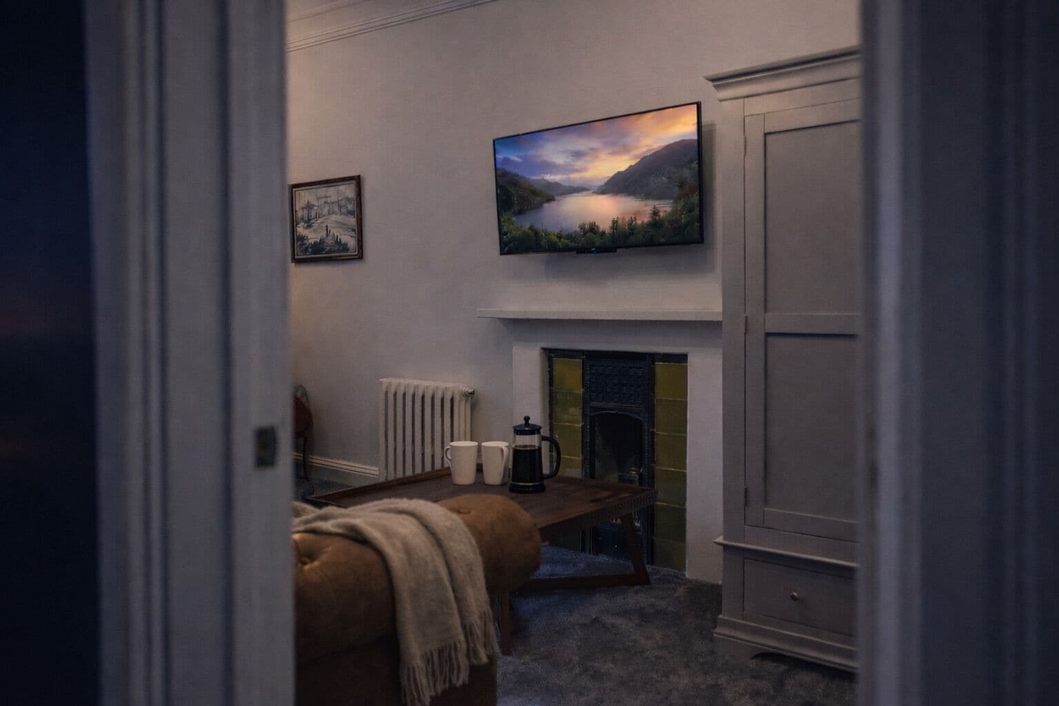 A cozy room with a fireplace, a bed, a table with a coffee pot, and a TV mounted above the fireplace.