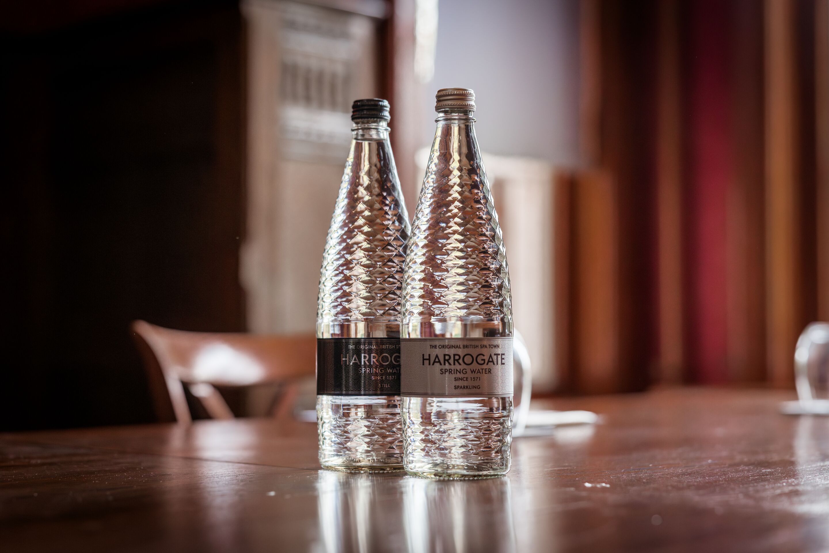 Zwei Harrogate Spring Water Flaschen auf einem Holztisch in einem Raum mit unscharfem Hintergrund.