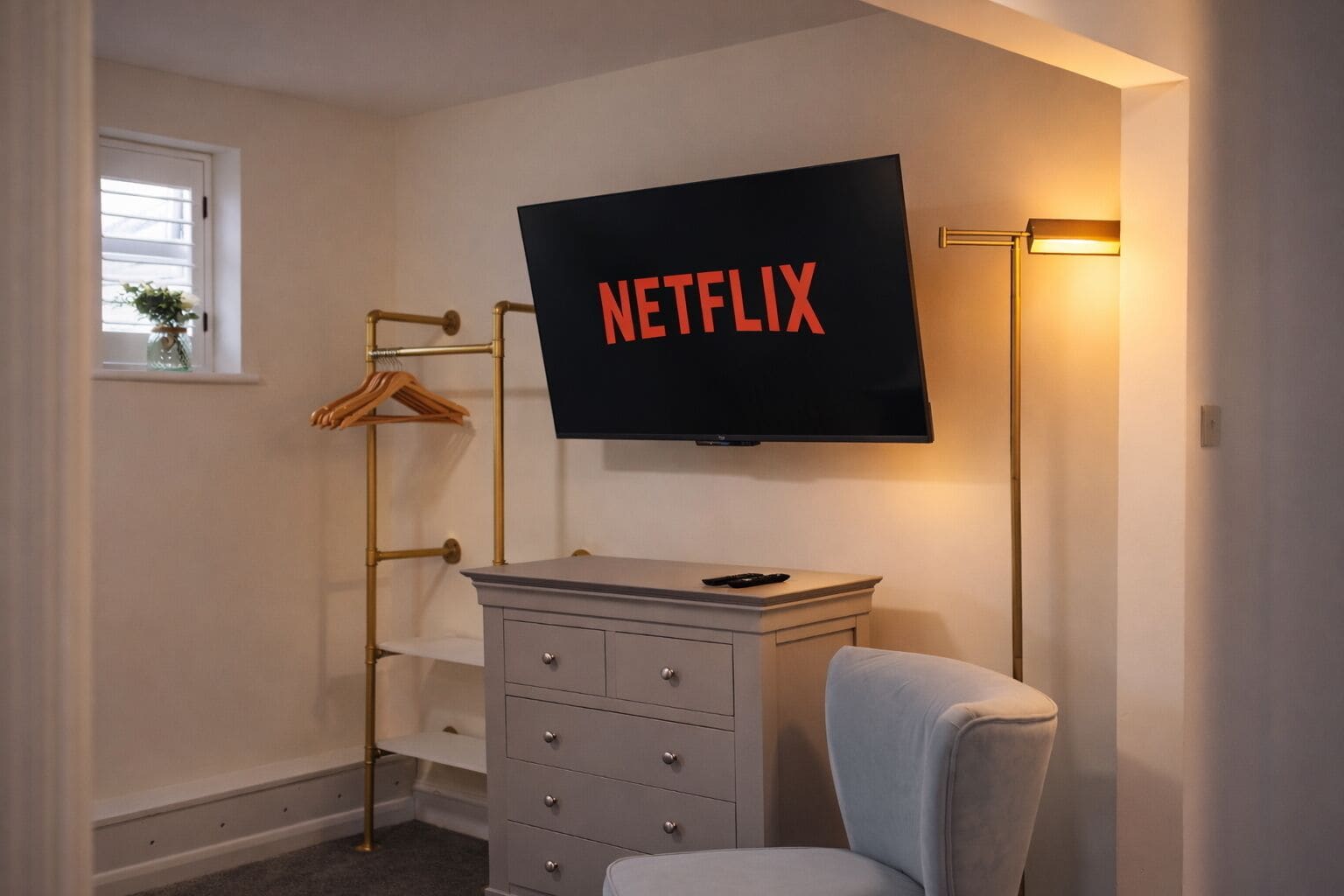 A bedroom with a TV mounted on the wall displaying the Netflix logo, a bedside table, a chair, and a wardrobe with a hanging rack.