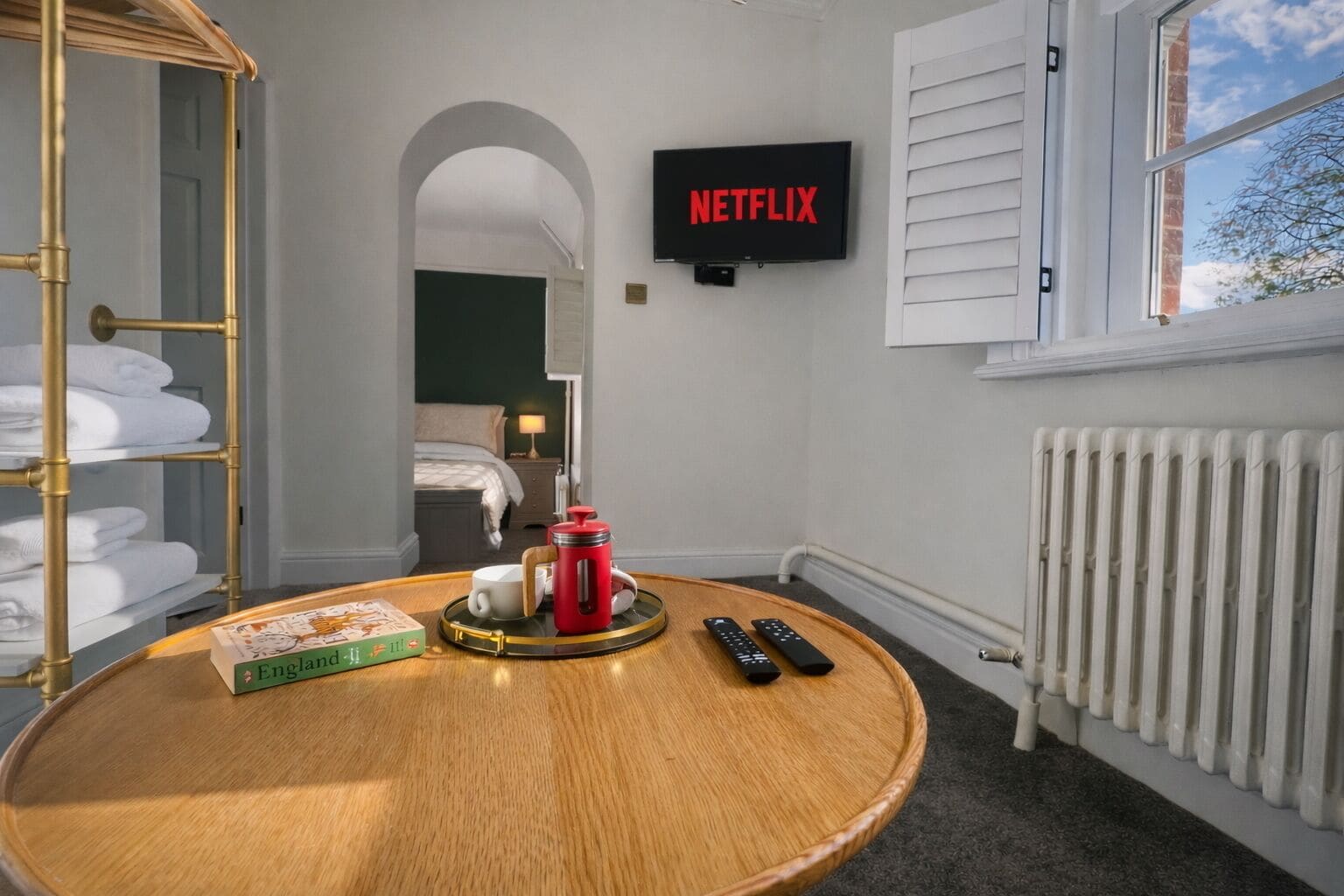 A hotel room with a round wooden table, a bookshelf, a bed, a radiator, and a TV displaying the Netflix logo.