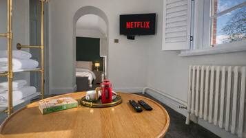 A hotel room with a round wooden table, a bookshelf, a bed, a radiator, and a TV displaying the Netflix logo.