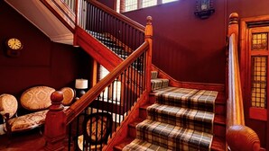 Un escalier en bois recouvert d’une moquette à carreaux, un lustre et une horloge au mur.