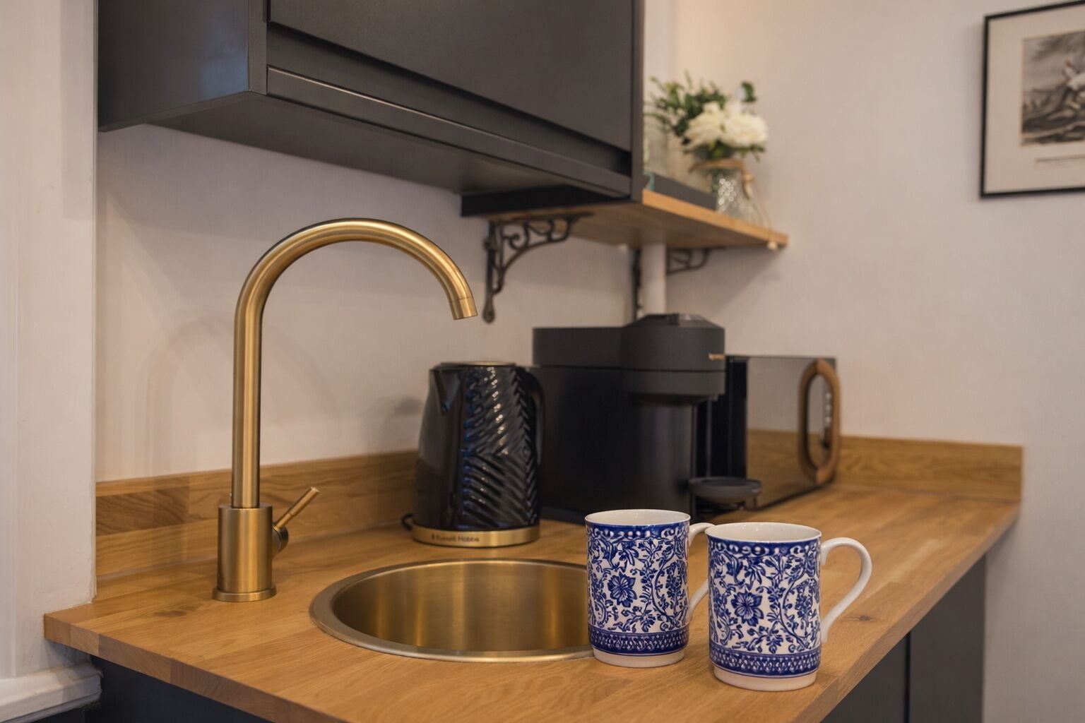 A kitchen counter with a gold faucet, a coffee maker, and two patterned mugs.