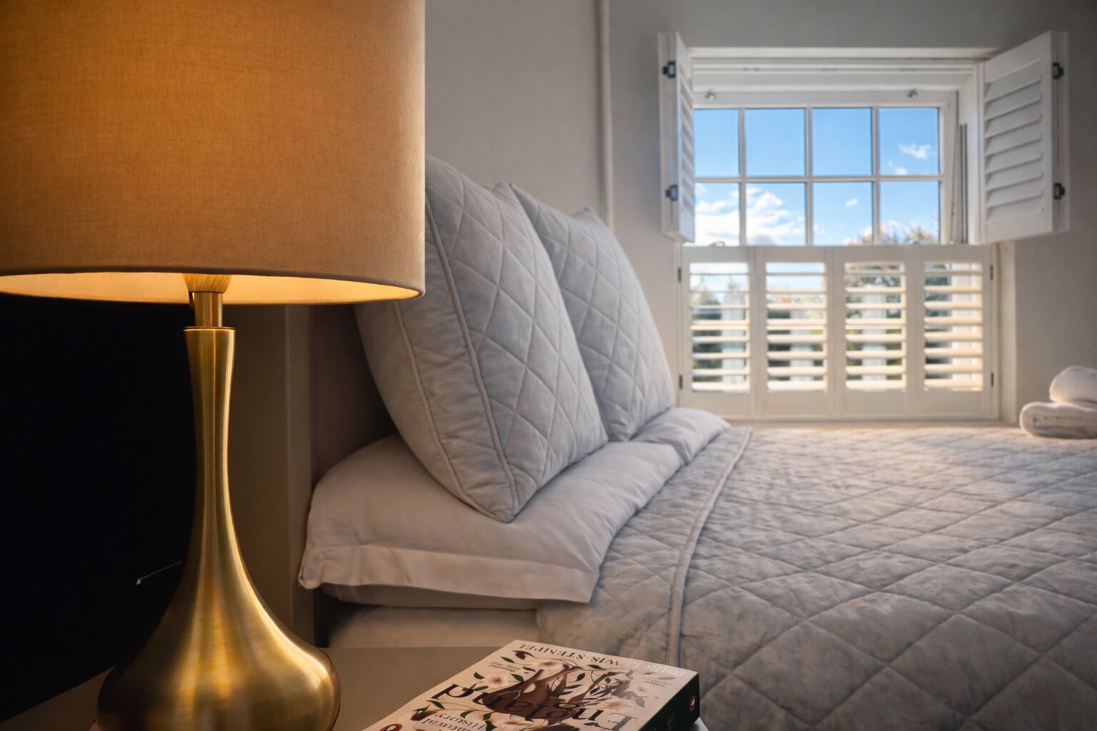 A bedroom with a bed, a bedside lamp, a window with shutters, and a book on the nightstand.