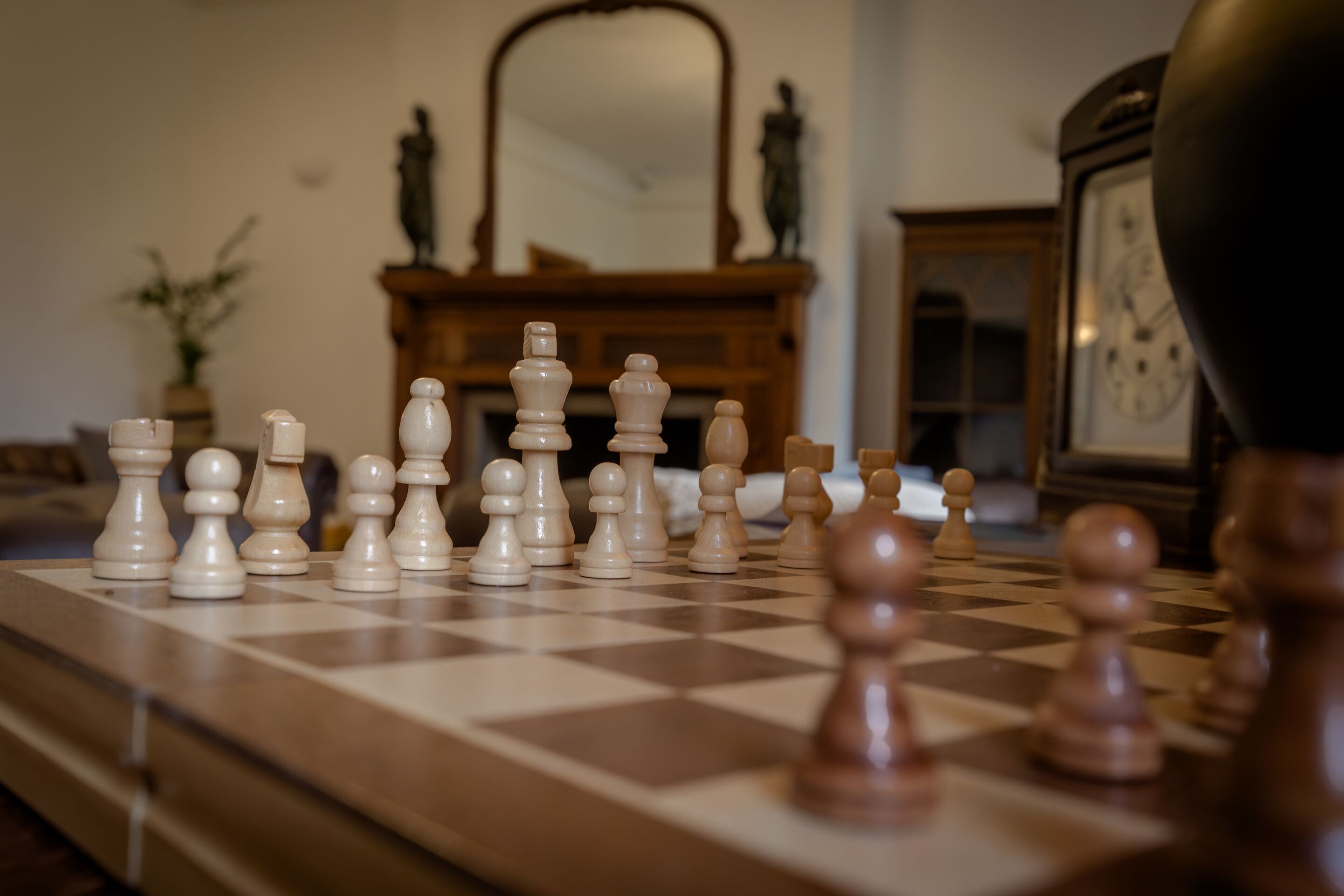 A chessboard with pieces set up in the center of a room with a mirror and a clock on the wall.