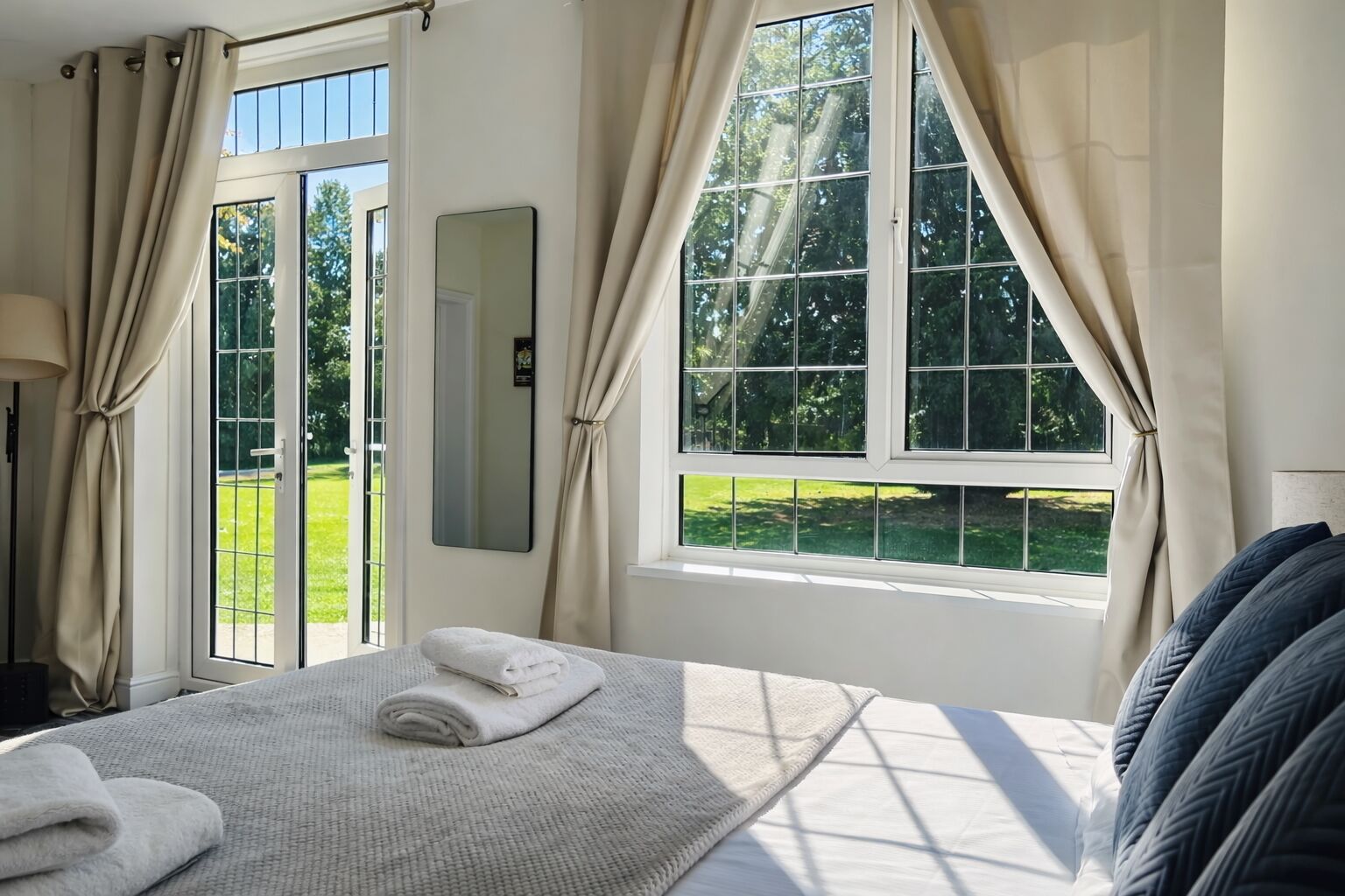A bedroom with a large bed, a window with curtains, a mirror, and a view of a green outdoor area.