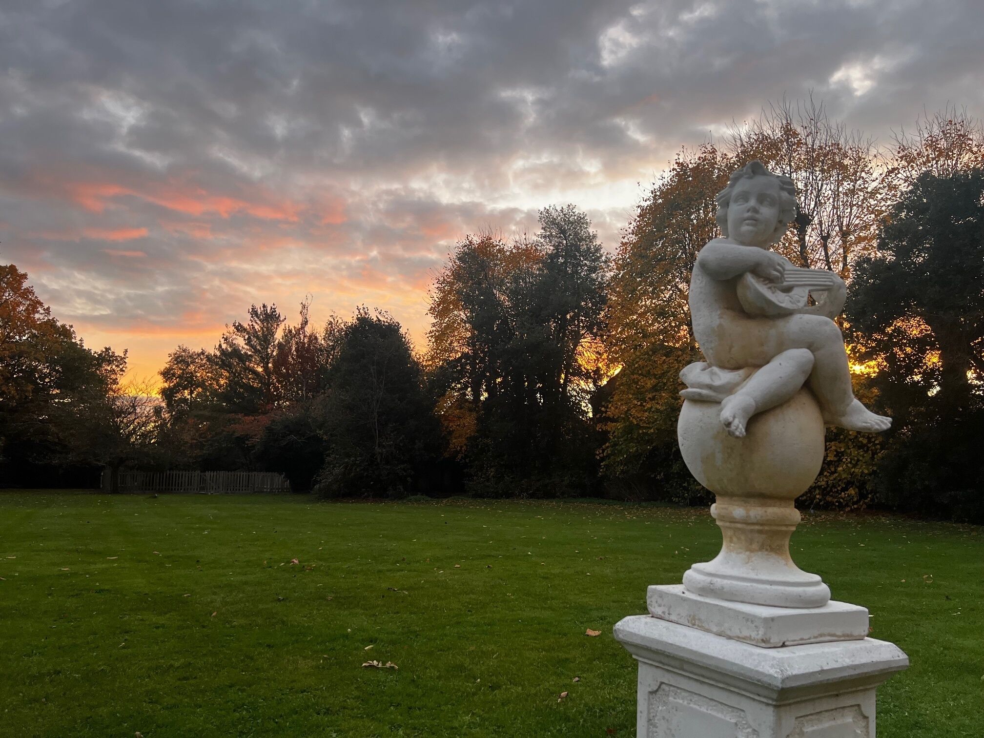 A statue of a seated figure on a pedestal in a grassy area with trees and a cloudy sky in the background.