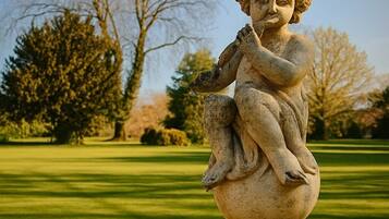 Una estatua de piedra de un ángel infantil sobre un pedestal en un parque con un campo cubierto de césped y árboles al fondo.