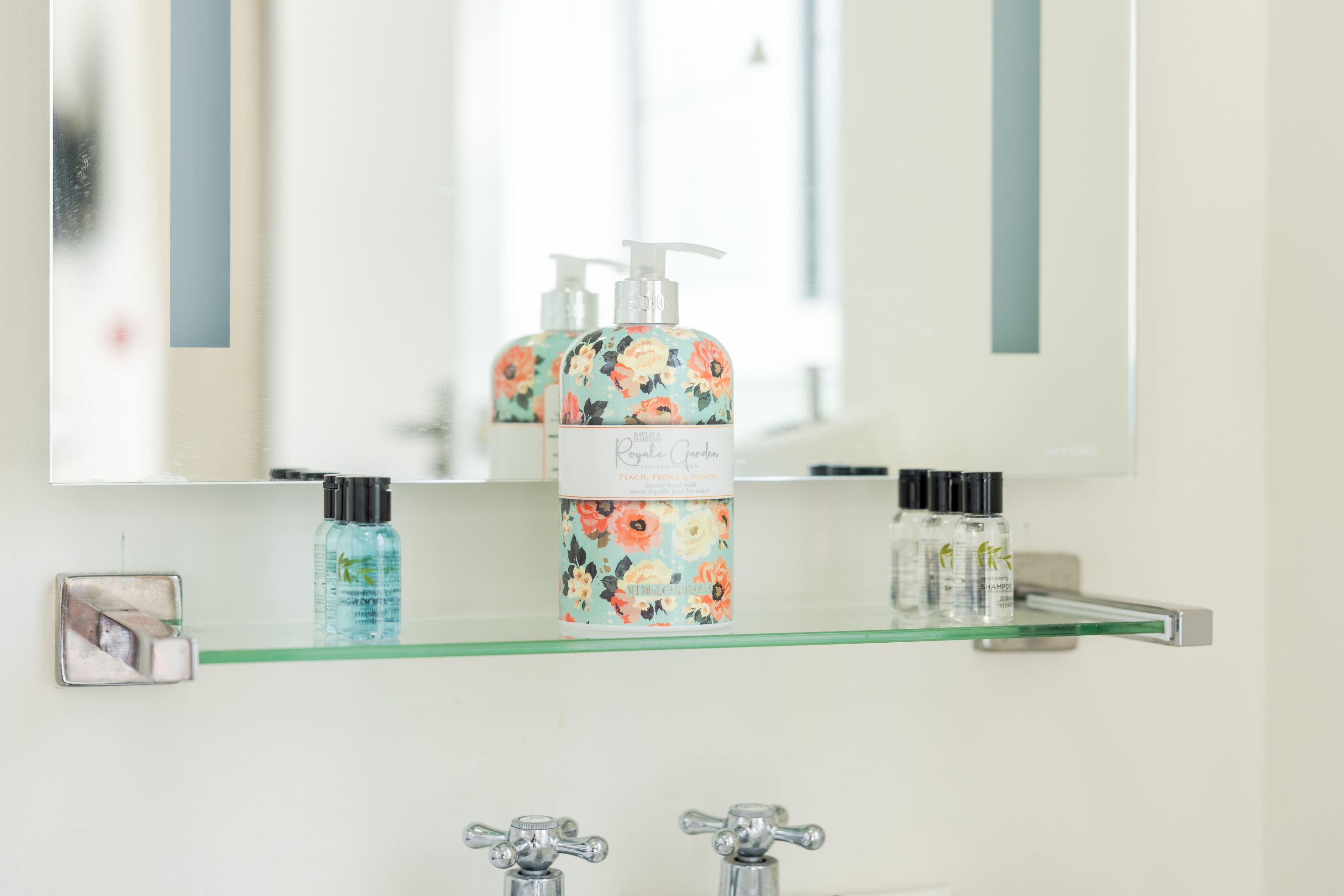 A bathroom with a glass shelf holding toiletries, a mirror, and two faucets.