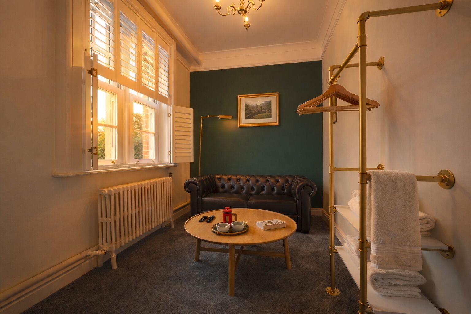 A cozy room with a black leather sofa, a round wooden table with a remote control, a book, and a cup. There's a radiator, a window with blinds, and a towel rack with towels.