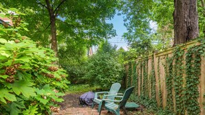 A brick pathway leading to a green Adirondack chair in a garden area with a wooden fence covered in ivy.