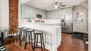 A modern kitchen with grey cabinets, a stainless steel refrigerator, a microwave, and a ceiling fan. There are three bar stools and a small table.
