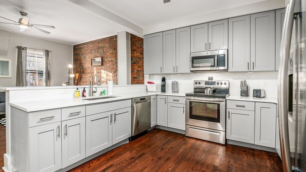 A modern kitchen with white cabinets, a brick accent wall, stainless steel appliances, and a wooden floor.