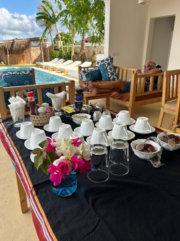 A table set with white cups, a vase with flowers, and various condiments, overlooking a pool area with lounge chairs and a palm tree. - TATU INN Boutique Hotel Watamu (Watamu)