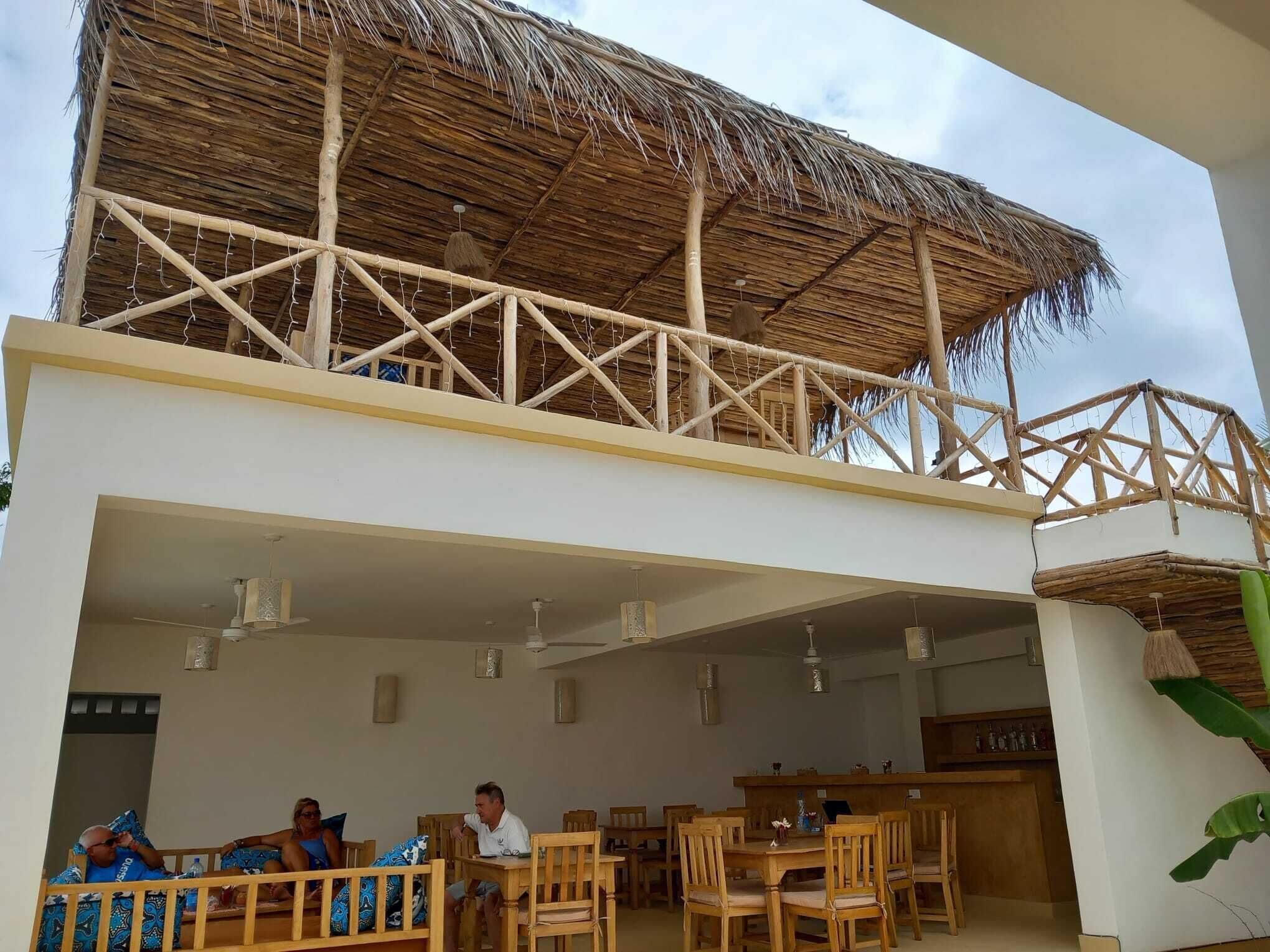 Un espace repas avec des tables et des chaises en bois, un bar et une structure dotée d’un toit de chaume.