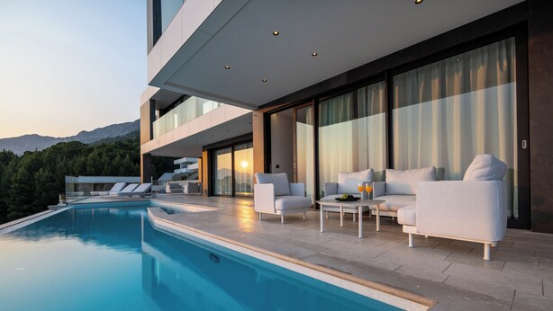 A modern outdoor pool area with lounge chairs and a table, surrounded by large windows and a mountain view. - Villa Brela-Rogac 3 (Baska Voda)