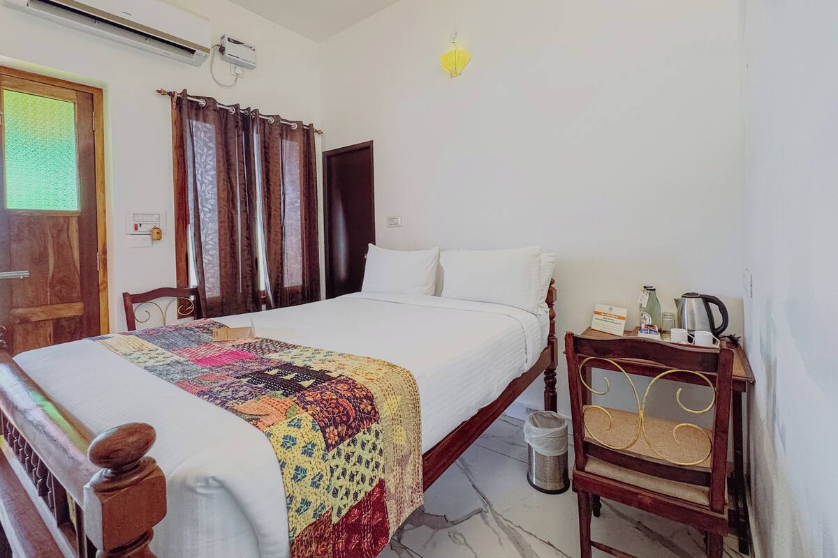 A hotel room with a bed, a bedside table with a kettle, a trash bin, and a window with curtains.