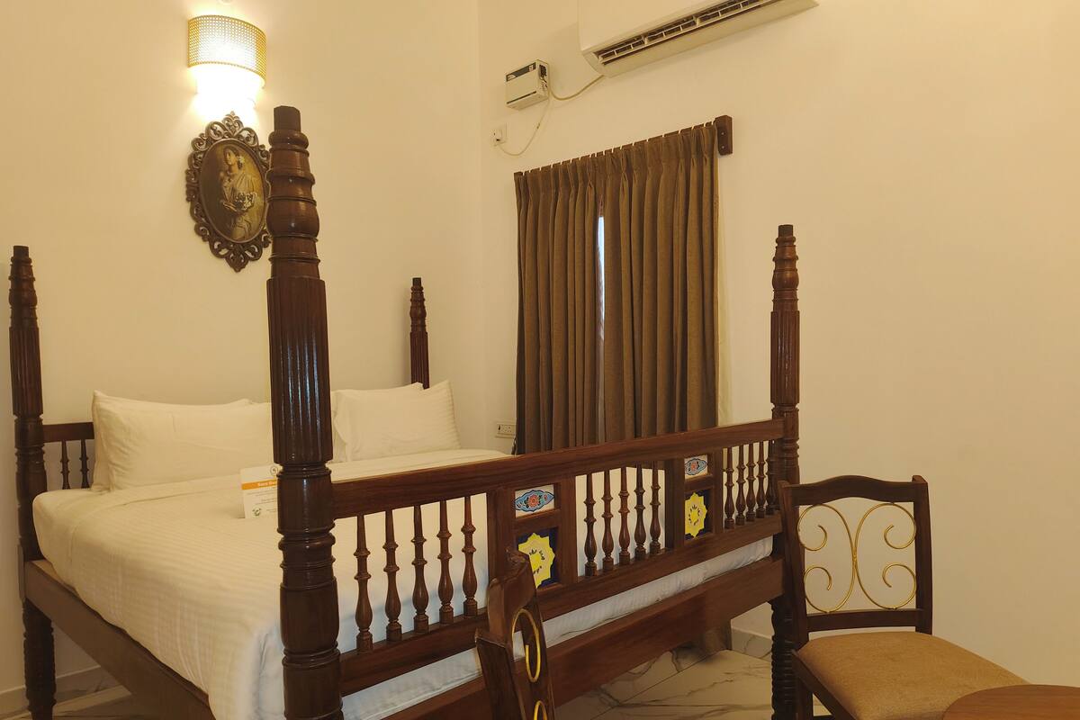 A four-poster bed with a wooden frame and white bedding, a small table, a chair, a wall-mounted air conditioner, and a wall-mounted light fixture.