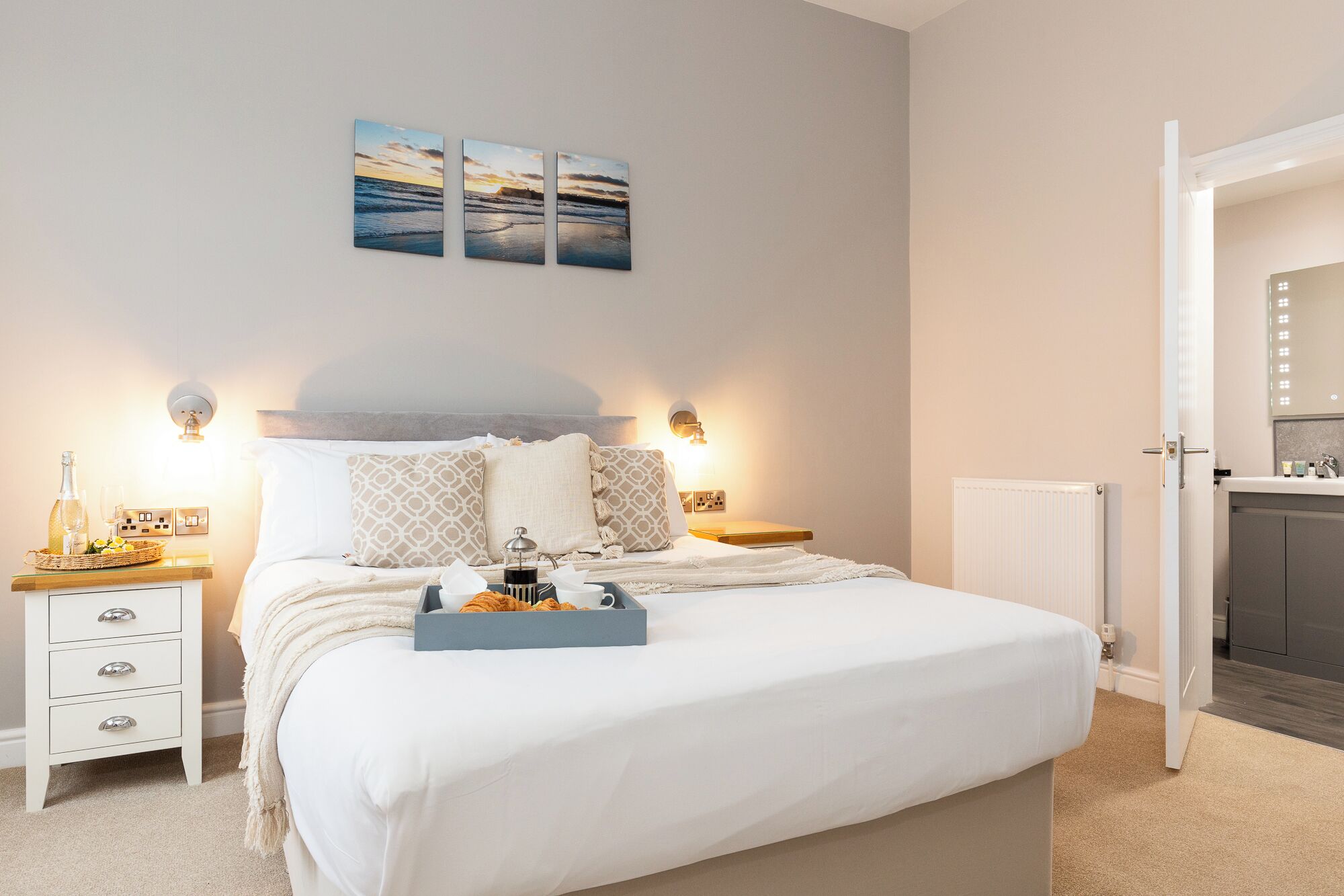 Une chambre bien rangée, avec un lit, des tables de chevet et une commode. On y trouve des lampes fixées au mur, un radiateur et une porte donnant sur une salle de bain.