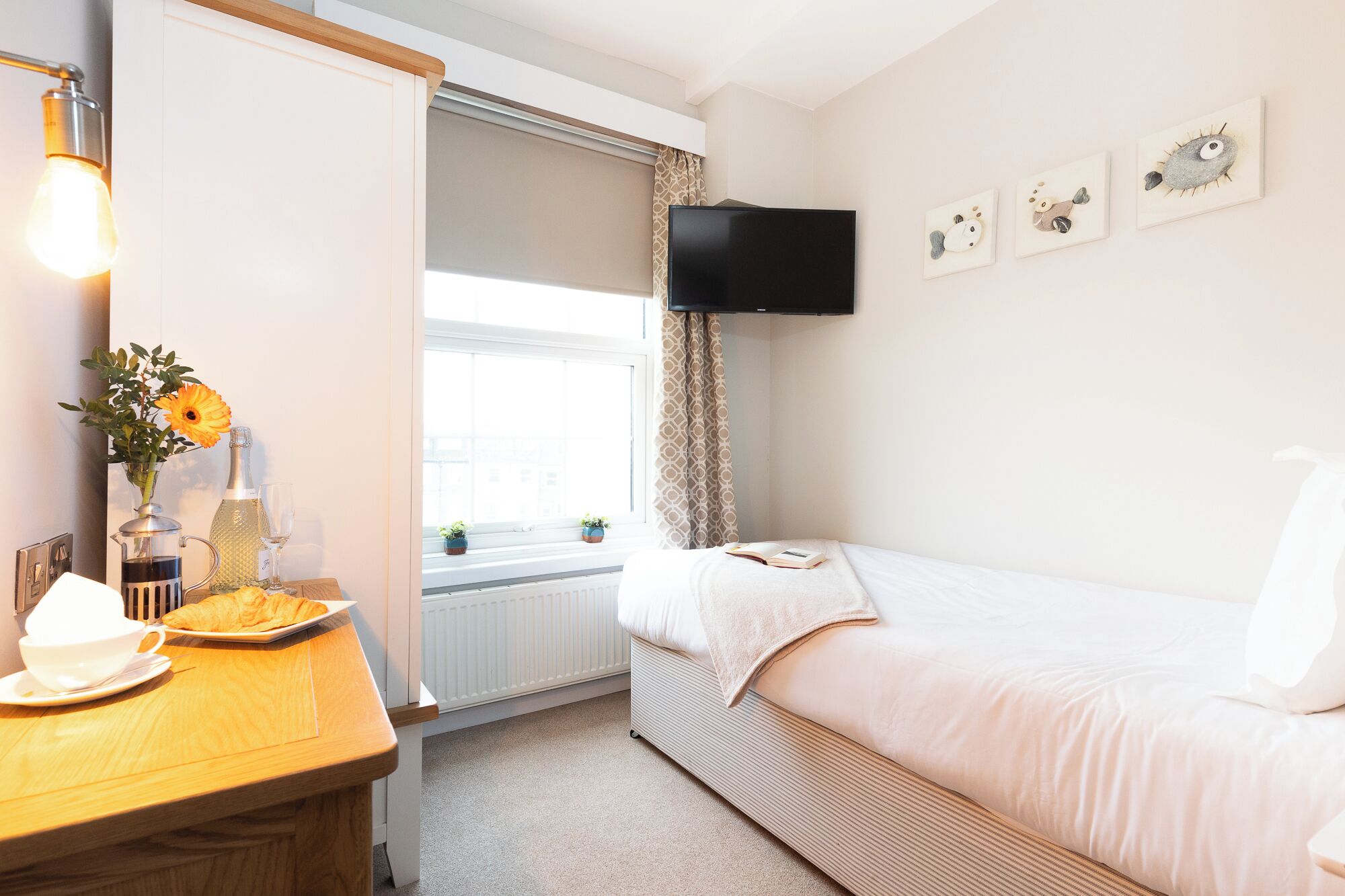 A bedroom with a bed, a bedside table with a lamp, a window with curtains, and a wall-mounted TV.