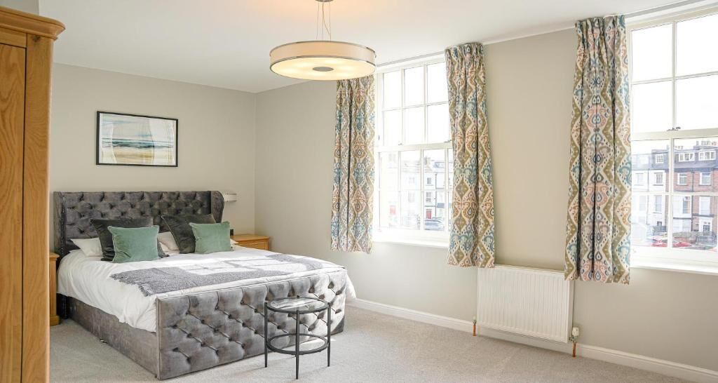 A bedroom with a tufted headboard bed, a small stool, patterned curtains, and a view of buildings through the window.