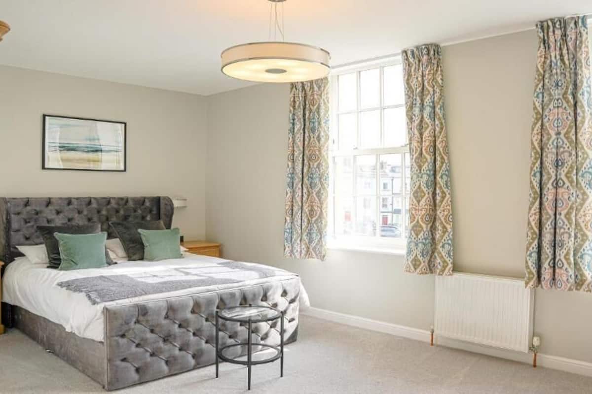 A bedroom with a tufted headboard bed, a small stool, patterned curtains, and a view of buildings through the window.
