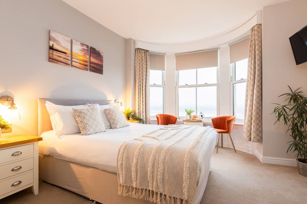 A bedroom with a large bed, a desk, two chairs, a TV, and a window with curtains.