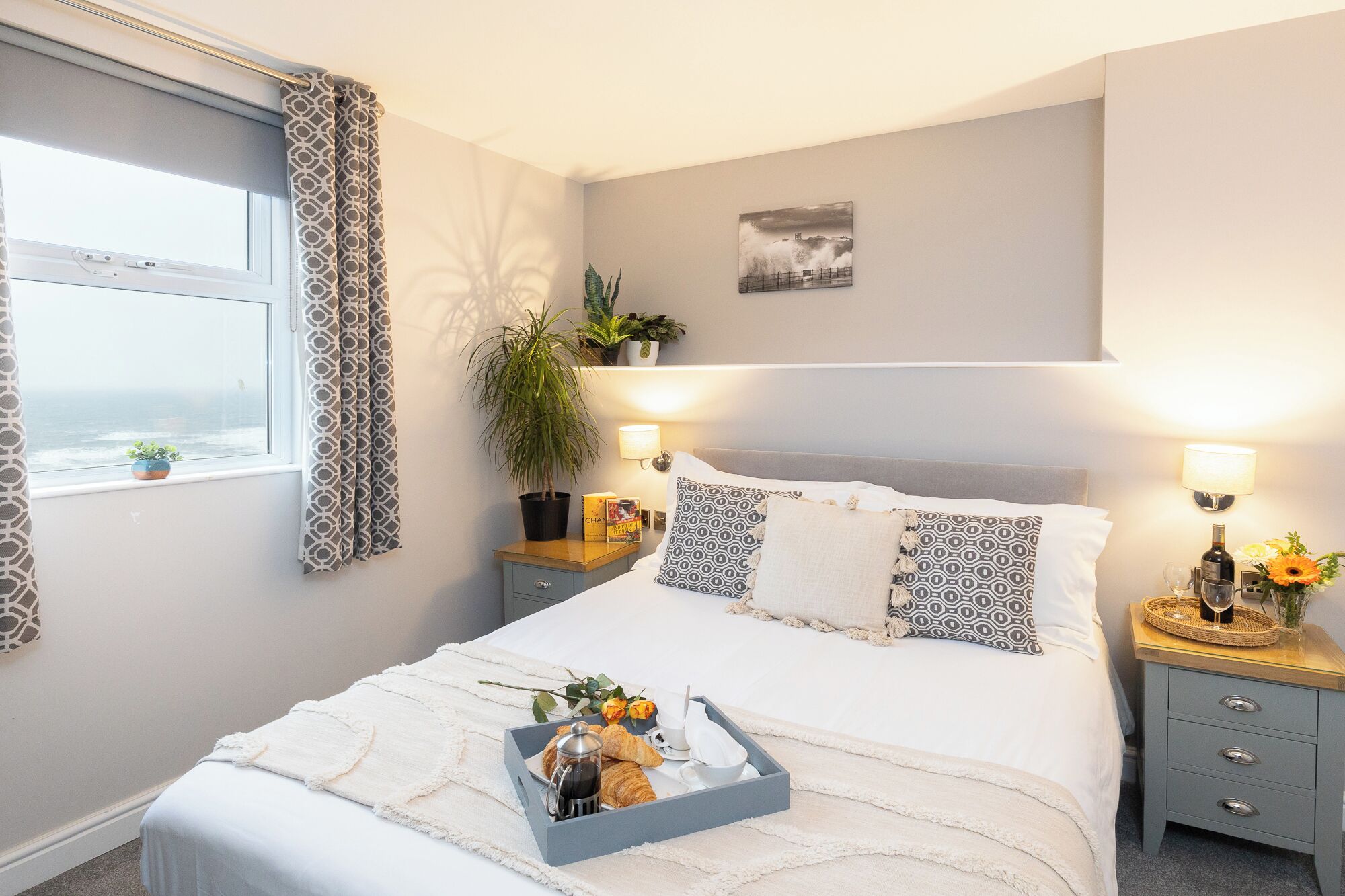 A bedroom with a bed, bedside tables, a window with curtains, a plant, a painting on the wall, and a tray with breakfast items.