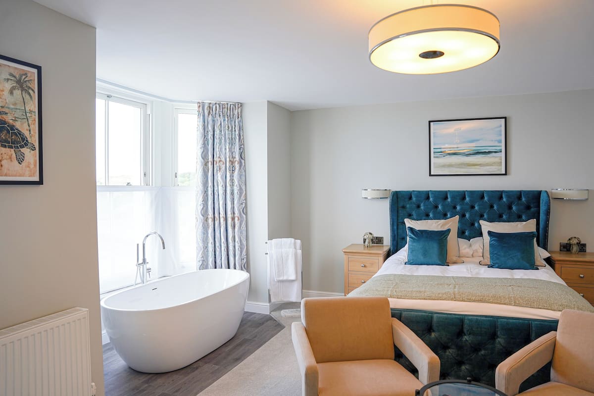 A modern hotel room with a large bathtub, a bed with blue headboard, and a view of the ocean through the window.
