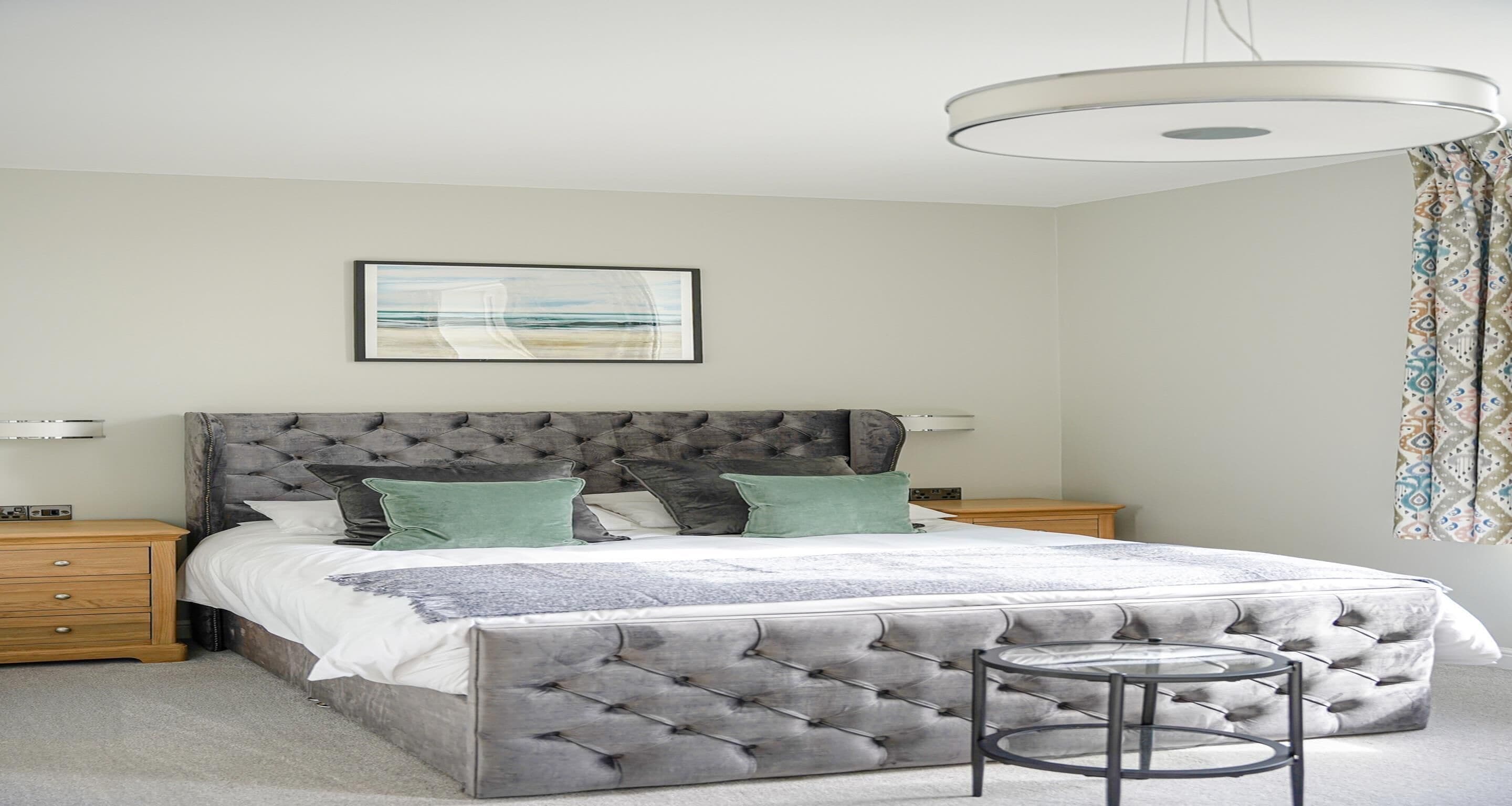 Un dormitorio con cabecera capitonada, cama con ropa de cama blanca, mesita de noche, una pequeña mesa redonda y una lámpara fijada a la pared.