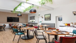 A modern restaurant with round tables and chairs, a large TV screen, and hanging plants. - The Clarence Gardens Hotel, BW Signature Collection (Scarborough)