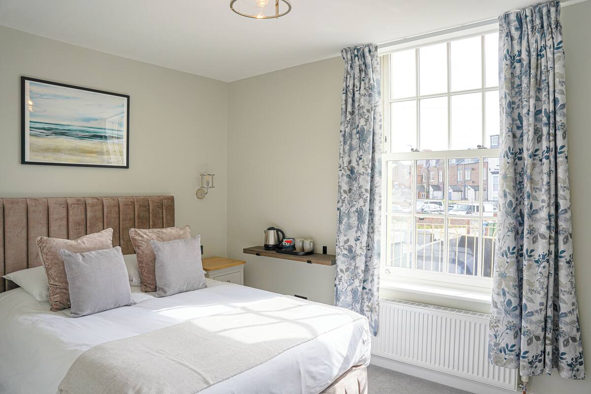 A bedroom with a bed, pillows, a nightstand, a window with curtains, a radiator, and a framed picture on the wall.