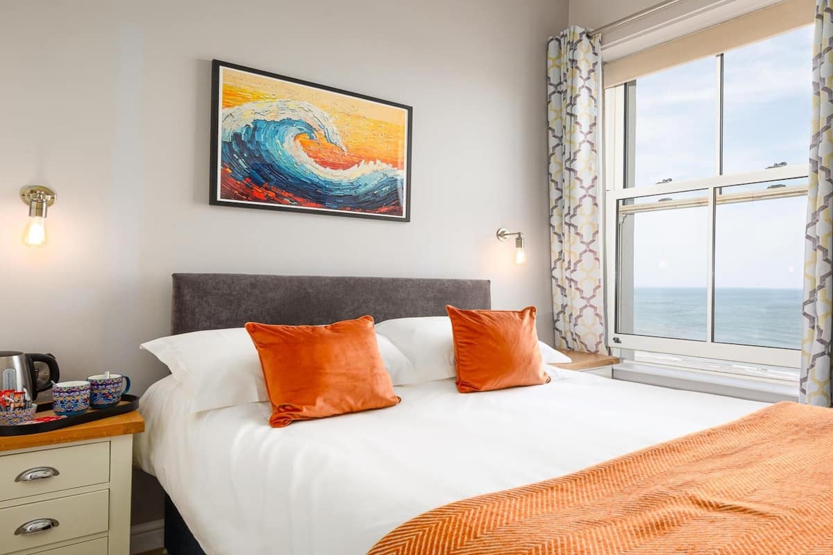 A hotel room with a bed, a painting, a window with curtains, and a nightstand with a kettle and cups.