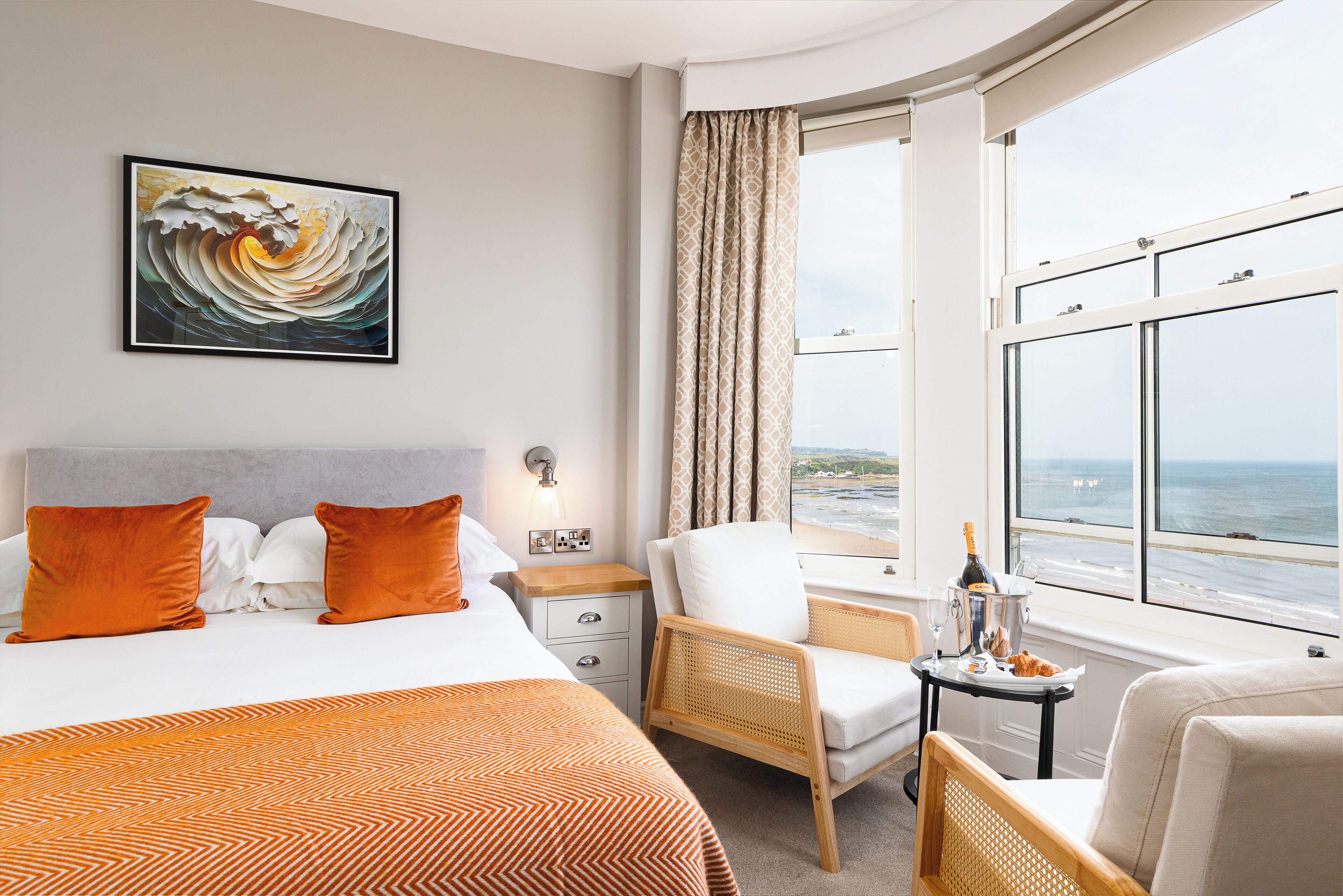 A hotel room with a bed, two chairs, a small table with a bottle and glasses, and a large window with a view of the sea.