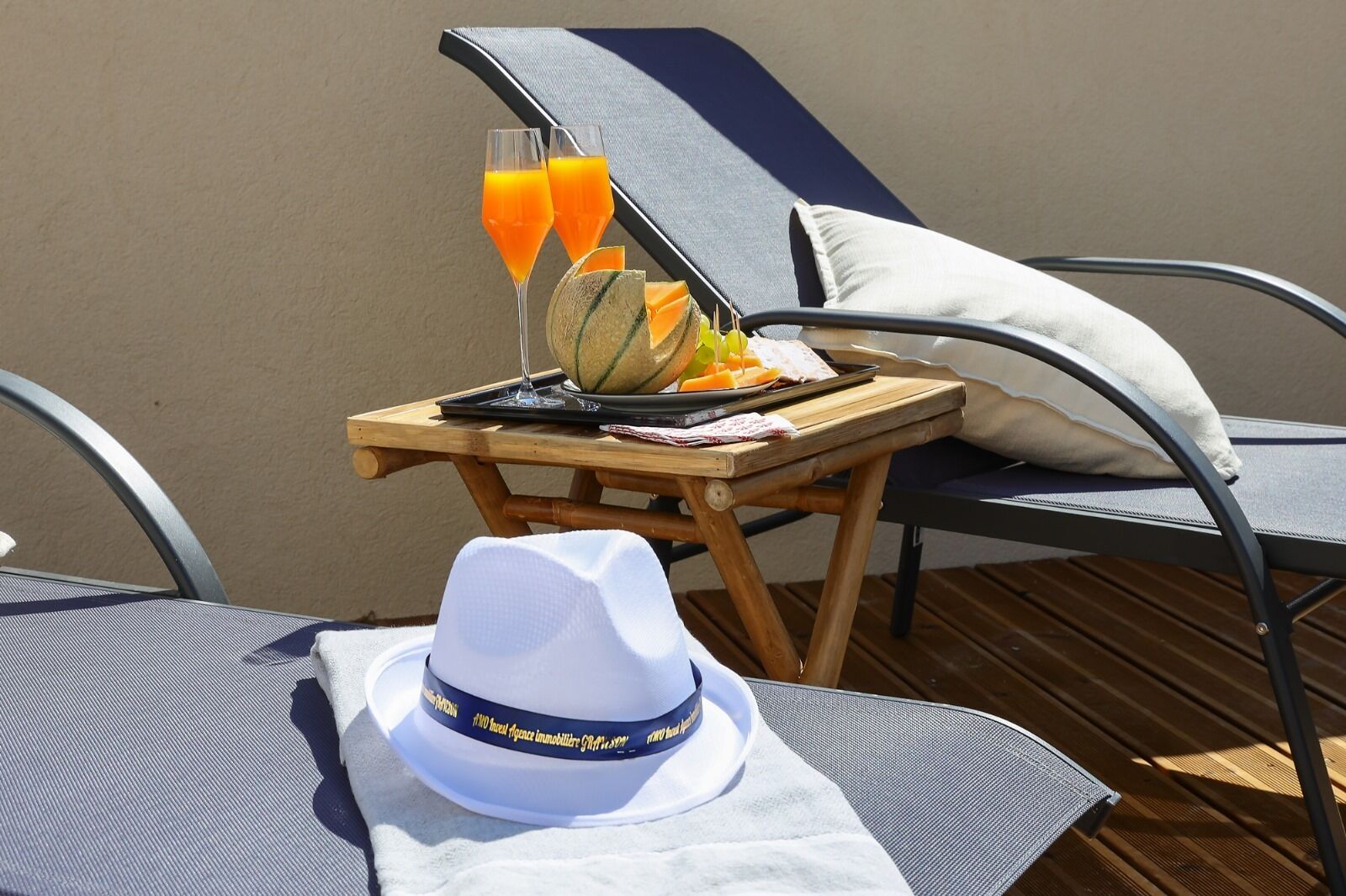 A sun lounger with a dining table beside it, a white hat, and a glass of orange juice.