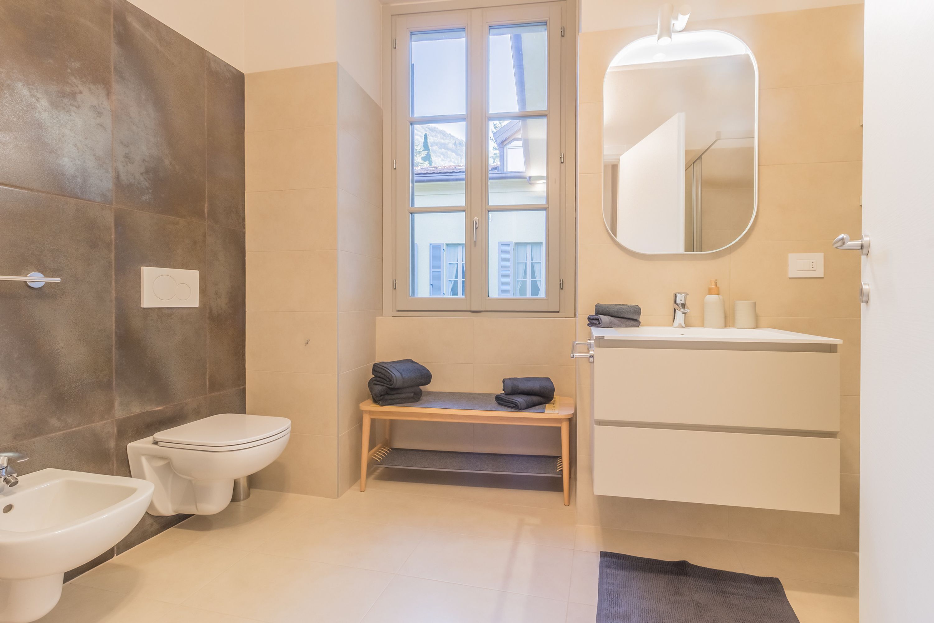 A bathroom with a toilet, bidet, and sink. There is a window with a view of buildings outside.