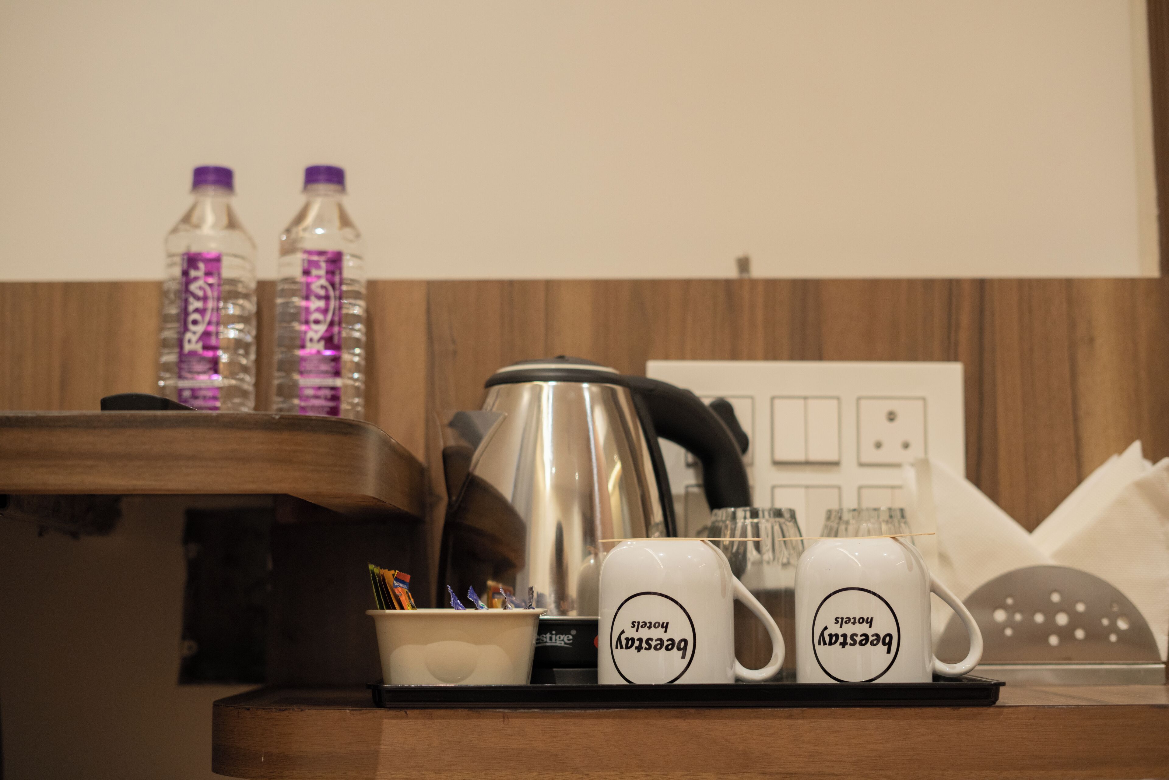A hotel room's minibar with two bottles of water, a coffee maker, and two mugs with the text "Beesly Roasters".
