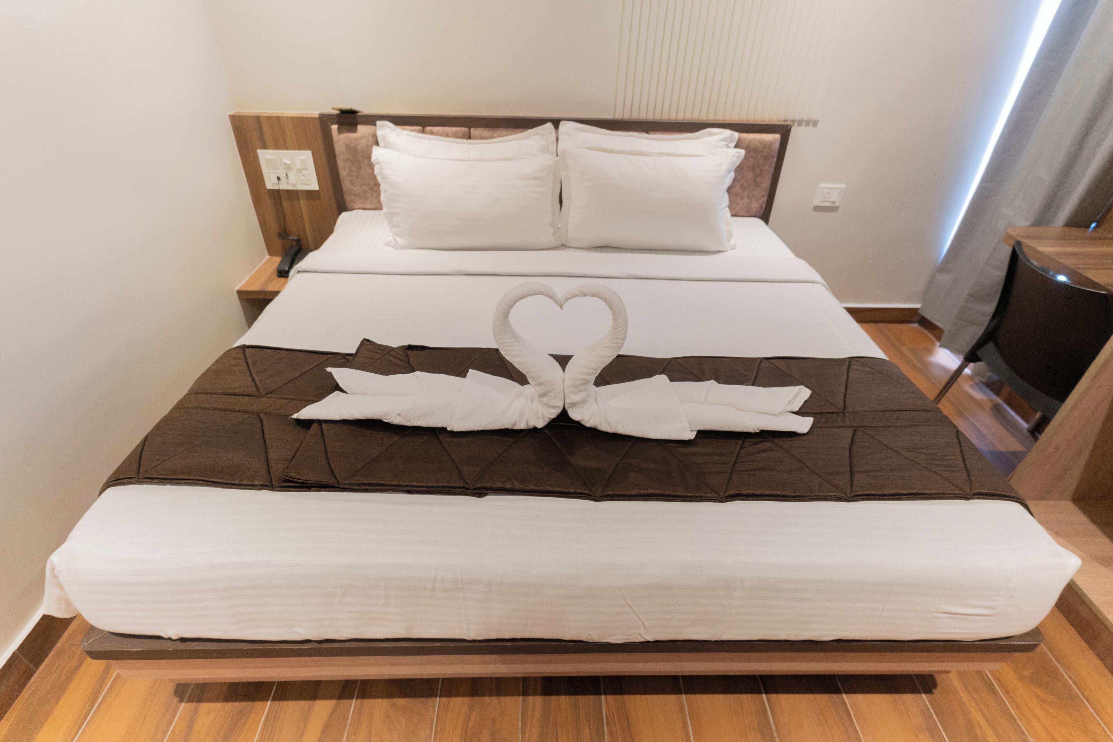 Un lit bien fait, agrémenté d’une serviette pliée en forme de cœur, avec une tête de lit en bois et une table de chevet sur laquelle se trouve une lampe.