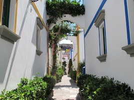 A narrow, white corridor with blue and yellow trim, flanked by lush green plants and a clear sky visible through an archway.