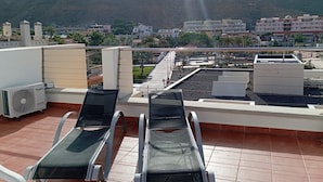 A rooftop terrace with two lounge chairs, a water fountain, and a view of a cityscape with buildings and a mountain in the background. - Playa de Mogán Alysio 8 (Mogan)