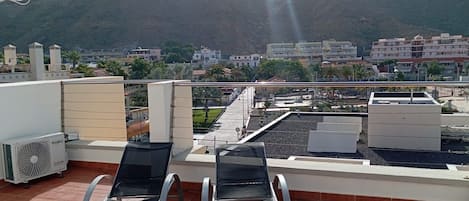 A rooftop terrace with two lounge chairs, a water fountain, and a view of a cityscape with buildings and a mountain in the background.