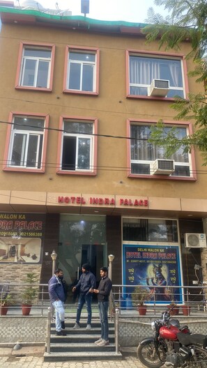 A building with multiple windows and a sign for Hotel Indra Palace. There are three people standing on the steps in front of the entrance. - Hotel Indra Palace By GRB (Vrindavan)